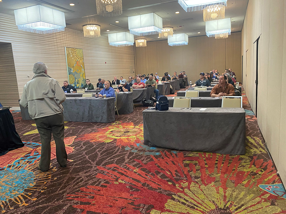 A presenter stands at the front of a conference room with attendees sitting at tables.