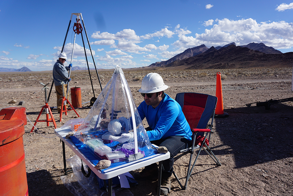 Researcher working at a table covered in a clear protective tent in the desert with mountains in the background.