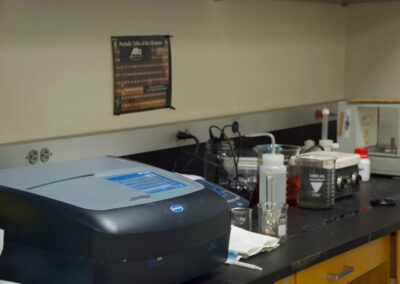 Beakers and various lab equipment is seen on a counter in a lab with the periodic table tacked to the wall.