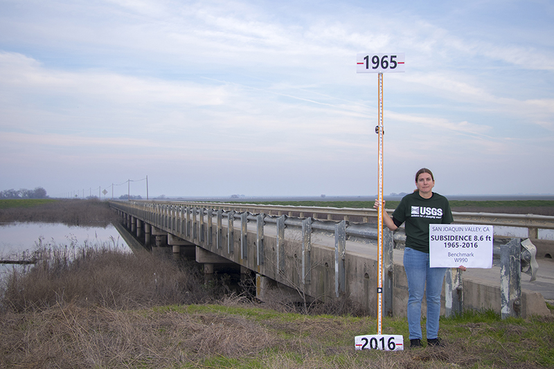 photo of researcher from USGS holding a measuring stick in front of a bridge.