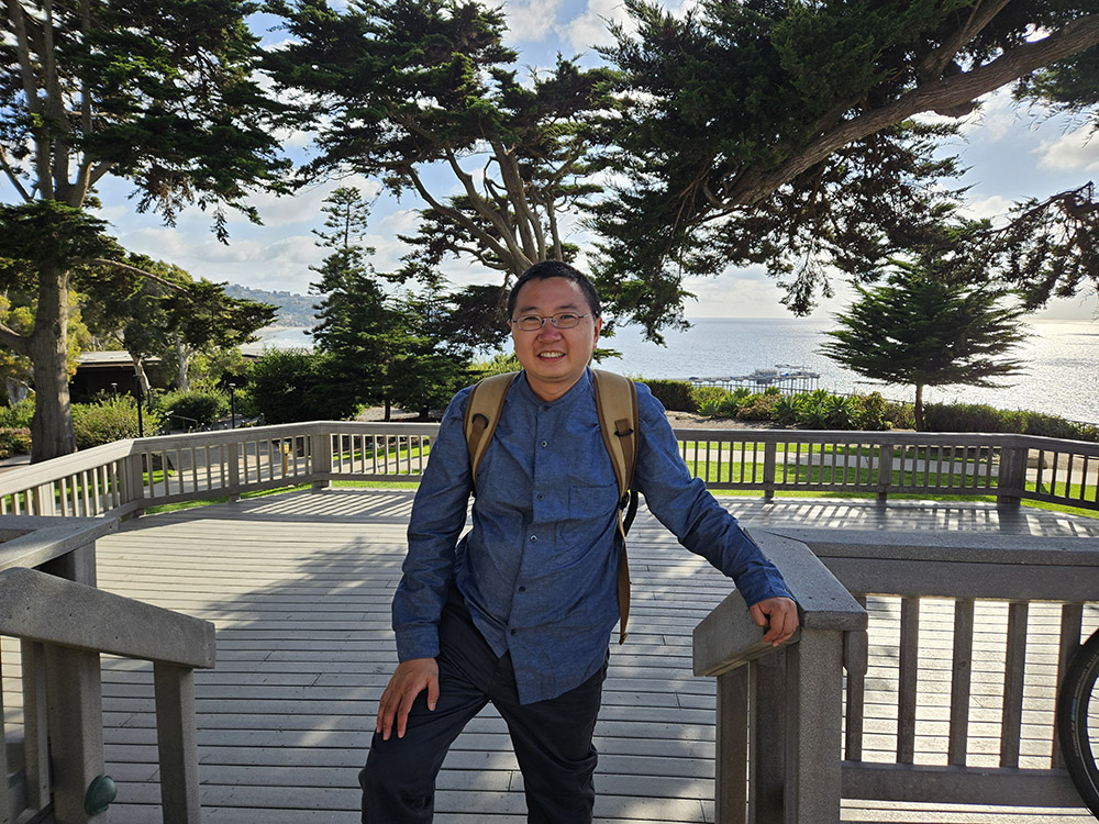 Ziming Ke standing on a deck under trees with the coast of a body of water in the background.