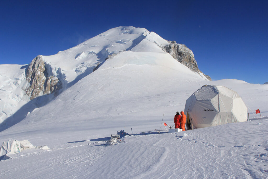 Two researchers in orange winter gear stand next to their tent on the side of a snowcapped mountain.